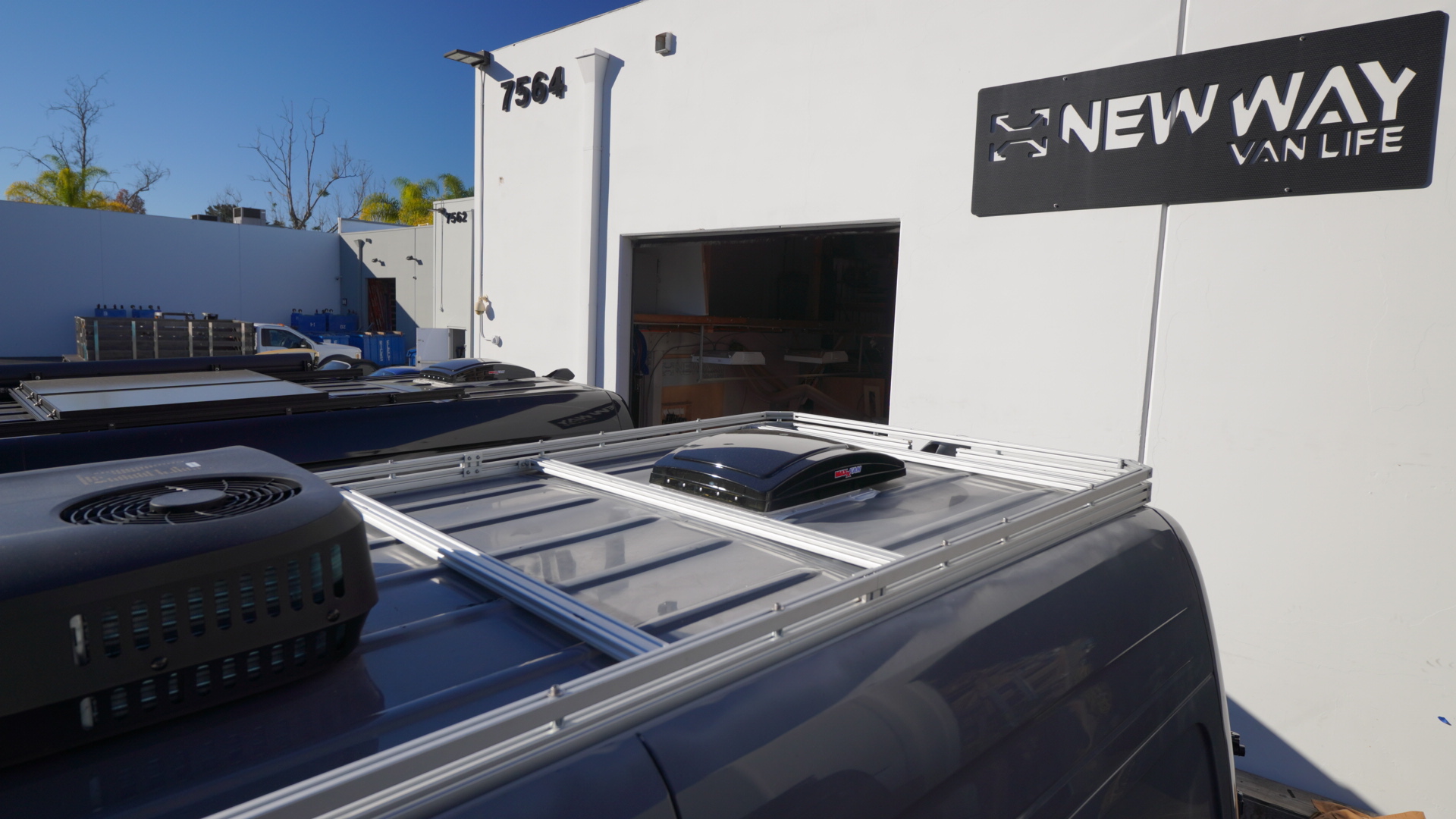 New Way Van Life silver aluminum roof rack installed on a 2021 Mercedes Sprinter van with a MaxxAir MaxxFan vent mounted on the platform, photographed outside the New Way Van Life facility in San Diego