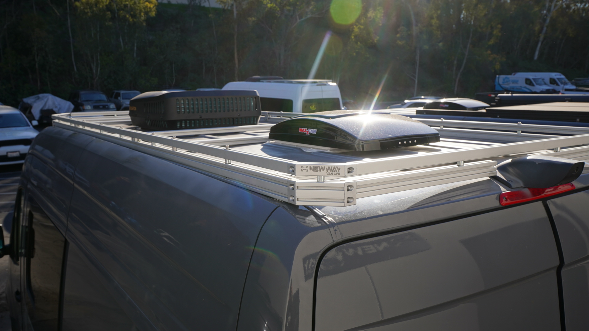 New Way Van Life silver aluminum roof rack installed on a 2022-2026 Mercedes Sprinter van with a MaxxAir MaxxFan roof vent and rooftop AC unit mounted on the platform shown from a rear three-quarter angle