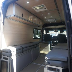 75" 3-door birch overhead cabinet installed in a Mercedes Sprinter van conversion showing closed cabinet doors, recessed ceiling lighting, seating area, and full van interior layout from the rear doors