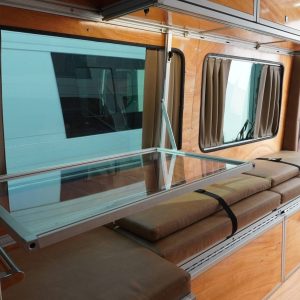 Full length interior view of Mercedes Sprinter camper van conversion with fold-away desk deployed, tan bench cushions with retention straps, warm wood walls, privacy curtains, overhead cabinets, and recessed ceiling light