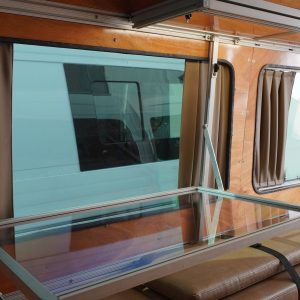 Mercedes Sprinter camper van conversion interior with fold-away desk fully deployed over tan bench seating, aluminum extrusion framing and warm wood panel walls visible alongside privacy curtains and overhead cabinets