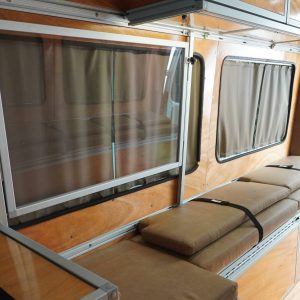 Mercedes Sprinter camper van conversion interior with fold-away desk in stored position showing full-length tan bench seating, privacy curtains, warm wood paneling, overhead cabinets, and under-bench storage drawers