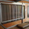 Mercedes Sprinter camper van conversion interior with fold-away desk stored and privacy curtains fully closed over side windows, tan bench seating with retention straps, warm wood panel walls, LED strip lighting, and overhead cabinets