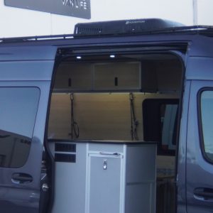 New Way Van Life Mercedes Sprinter camper van exterior evening view with sliding door open showing galley module, pull-out base storage drawer, and Houghton rooftop air conditioner under the New Way Van Life sign