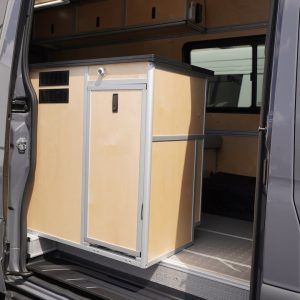 Close-up view through open sliding door of a New Way Van Life Mercedes Sprinter conversion showing the birch plywood galley module with locking cabinet door, ventilation grilles, and aluminum extrusion framing against the van interior and overhead cabinets