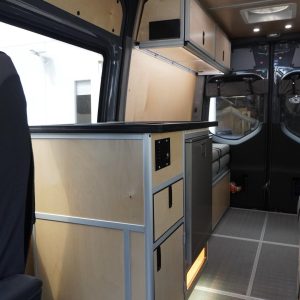 Interior view of New Way Van Life Mercedes Sprinter conversion showing the full galley layout with dark countertop, stacked plywood drawers, overhead storage cabinets with LED under-lighting, floor-level amber accent lighting, and ceiling-mounted air conditioning unit