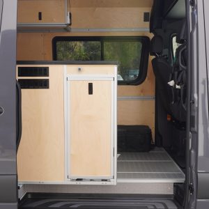 Interior of New Way Van Life Mercedes Sprinter conversion viewed through open sliding door showing birch plywood galley module, overhead storage cabinets, recessed LED ceiling lights, and ceiling-mounted air conditioning unit with trees visible through the rear partition window