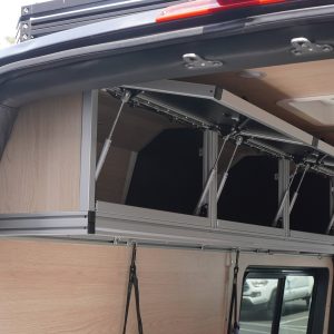 3-door birch overhead cabinet with all lift-assisted doors fully open showing gas strut hardware viewed from the rear door opening of a Mercedes Sprinter van conversion by New Way Van Life