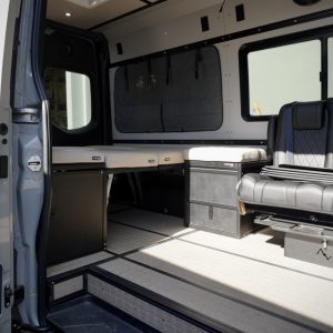 Custom gray and cream automotive-grade vinyl bed system cushions installed on driver side platform inside a Mercedes Sprinter van conversion by New Way Van Life, shown from the sliding door entrance