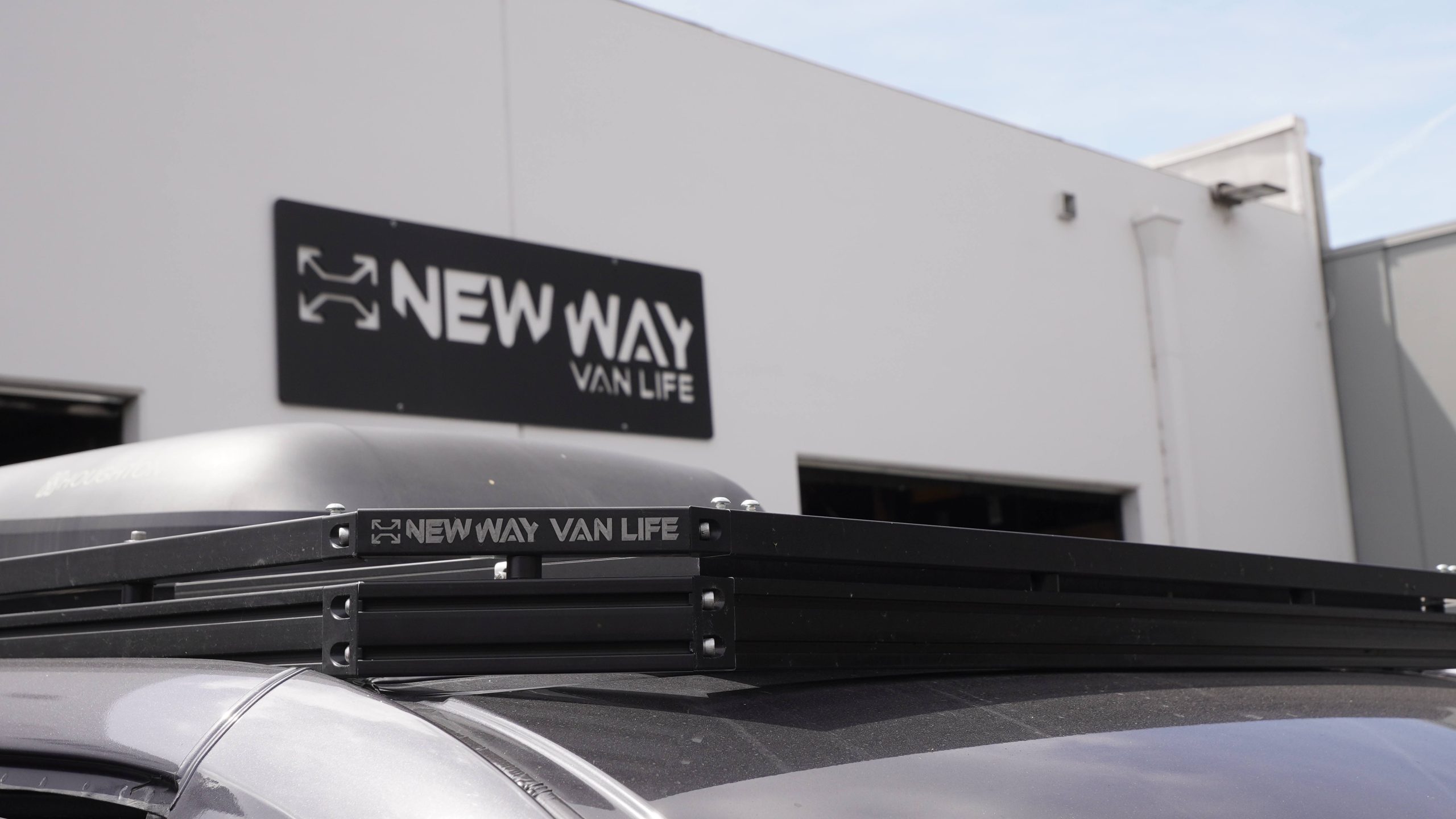 New Way Van Life branded black aluminum roof rack on a Mercedes Sprinter van with Houghton AC unit mounted on platform and New Way Van Life shop sign prominently visible in background