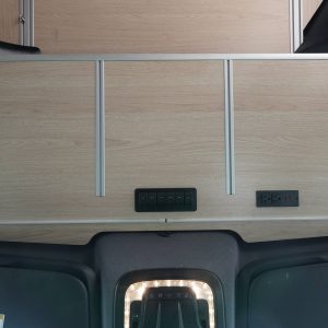 Sprinter van cockpit upper shelf deployed above the cab showing birch laminate surface, aluminum extrusion frame, integrated rocker switch panel, and 110V and USB power outlets