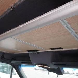 Sprinter van cockpit upper shelf shown from below at an angle highlighting the birch laminate underside, aluminum extrusion frame rails, and quick-release mounting hardware above the cab area