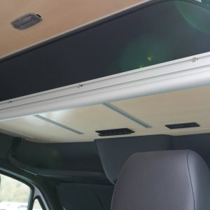 Sprinter van cockpit upper shelf shown in deployed position from the passenger side angle highlighting the birch laminate surface, aluminum extrusion frame, black fabric ceiling panel, and integrated power modules above the cab area