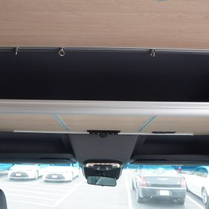 Sprinter van cockpit upper shelf shown in stored position against the ceiling with black fabric panel facing down, aluminum extrusion frame, and curtain ring hardware visible above the cab area