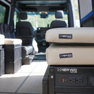 Two stacked cream automotive-grade vinyl bed system cushions with New Way Van Life branded labels sitting on top of a New Way Van Life branded power module inside a Mercedes Sprinter van conversion showing the full dual platform bed system and ceiling AC unit in the background