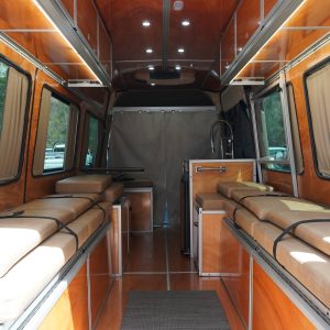 Full interior view of a Mercedes Sprinter van conversion with cherry wood panels dual tan cushion bed platforms amber LED lighting overhead cabinets and open window curtains by New Way Van Life