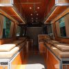 Rear door view looking into a fully built Mercedes Sprinter van conversion with cherry wood panels dual tan cushion bed platforms LED lighting power modules and kitchen unit visible in the background by New Way Van Life