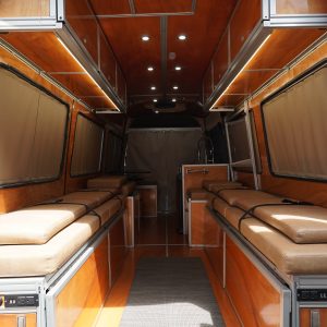 Rear door view looking into a fully built Mercedes Sprinter van conversion with cherry wood panels dual tan cushion bed platforms LED strip lighting and window curtains closed by New Way Van Life