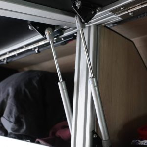 Interior view of a Sprinter van overhead cabinet fully open with dual gas strut lift assist arms extended showing stored gear inside the cabinet and van interior in the background by New Way Van Life