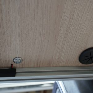 Interior underside of a Sprinter van overhead cabinet showing a push button light switch and circular cable management grommet on birch laminate panel with aluminum extrusion frame by New Way Van Life