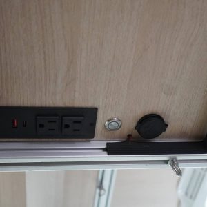 Interior underside of a Sprinter van overhead cabinet showing USB USB-C and dual 110V power outlets alongside a 12V port and push button switch on birch laminate panel by New Way Van Life